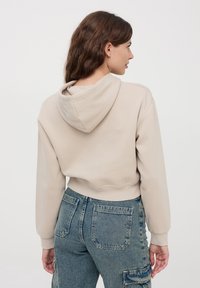 Krótkotali beżowy hoodie wykonany z miękkiego materiału. Posiada kaptur, ściągacze przy mankietach oraz surowe wykończenie u dołu. Zestawiony z niebieskimi dżinsowymi spodniami cargo w wysokim stanem.