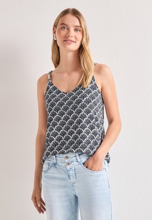 Młoda kobieta z długimi blond włosami, ubrana w czarno-biały wzorzysty top bez rękawów i jasnoniebieskie dżinsy, stoi z jedną ręką w kieszeni.