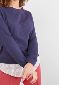 Granatowa bluza z długimi rękawami, okrągłym dekoltem oraz teksturowaną, jasną warstwą pod spodem, zestawiona z czerwonymi spodniami.