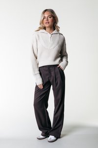 Suéter de punto grueso de color crema con un cuello de media cremallera, combinado con pantalones de cuadros marrón oscuro de corte suelto y zapatillas blancas con detalles en gris.