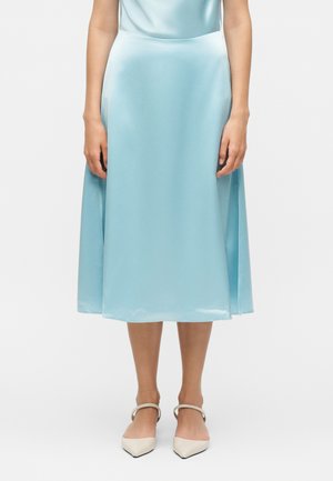 Woman standing wearing a light blue satin midi skirt and beige pointed-toe ankle-strap heels against a plain white background.