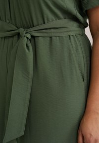 Maslinasto zelena haljina od teksturirane tkanine, s remenom u struku, dugmadi za zatvaranje i bočnim džepovima za dodatnu funkcionalnost.