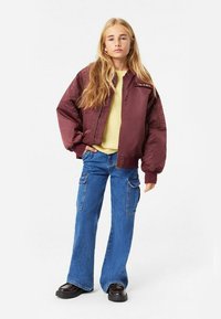 Burgunderfärgad bomberjacka med dragkedja fram, beige tröja och blå utsvängda carg Jeans. Svarta plattformsskor, långt blont hår, neutral bakgrund.