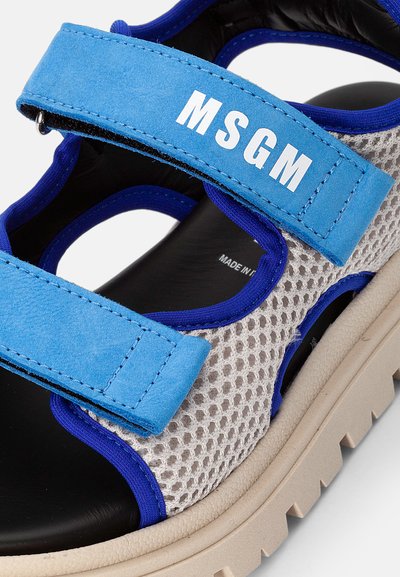 Sandalha em malha azul e cinza com tiras ajustáveis em Velcro. Apresenta uma sola grossa em bege e um forro preto. Nome da marca "MSGM" exibido.