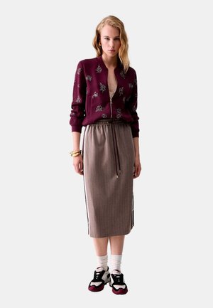 Donna con giacca bordeaux floreale con cerniera, gonna beige al ginocchio con coulisse, calzini bianchi e sneakers nere e bordeaux.
