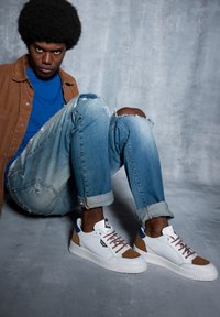Zapatillas deportivas blancas y marrones con acentos azules, punta redonda, suela texturizada y cordones marrones. Llevadas con vaqueros azules desgastados y una camisa azul.