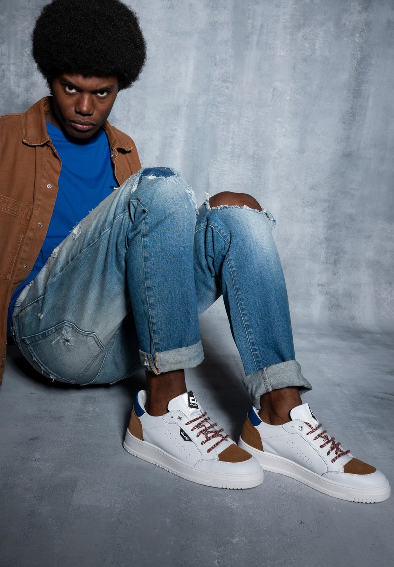 Zapatillas deportivas blancas y marrones con acentos azules, punta redonda, suela texturizada y cordones marrones. Llevadas con vaqueros azules desgastados y una camisa azul.