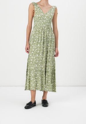 Vrouw die een mouwloze groene midi-jurk met bloemenprint draagt, met een V-hals en strikbandjes, gecombineerd met zwarte loafers, staand tegen een witte achtergrond.