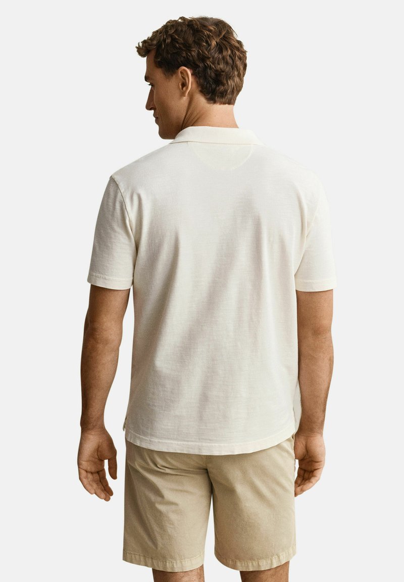 Mann, der ein weißes kurzärmeliges Poloshirt und beige Shorts trägt, steht mit dem Rücken zur Kamera vor einem schlichten Hintergrund.
