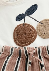 Bílé tričko s hnědým a béžovým chlupatým výšivkou třešní; hnědobílé pruhované šortky s elastickým pasem.