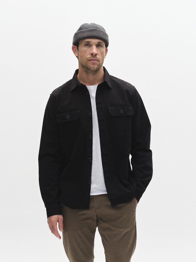 Juodas mygtukais užsegamas švarkelis su dviem krūtinės kišenėmis, dėvimas ant baltos marškinėlių. Modelis vilki pilką megztą kepuraitę ir rudos spalvos kelnes.