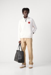 White zip-up sweatshirt with a red "Hugo" logo, beige trousers, and a black leather tote bag. Model wears white sneakers with gum soles.