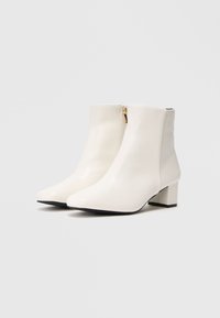 Bottines blanches en cuir lisse, avec un bout pointu et un petit talon carré. Fermeture éclair sur le côté avec des détails en métal doré.