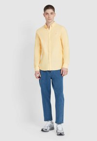 Geel knoopshirt van lichtgewicht stof, lange mouwen, enkele borstzak, gecombineerd met blauwe spijkerbroek en grijze sneakers.