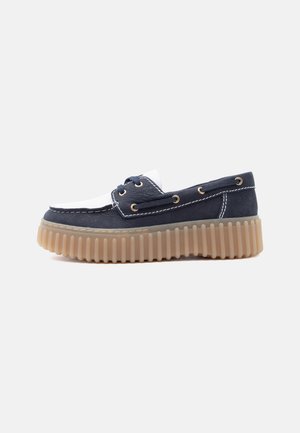 Scarpa da barca piattaforma in pelle blu navy e bianca con chiusura frontale a lacci, cuciture a contrasto e suola spessa in gomma scanalata color nocciola su sfondo neutro.