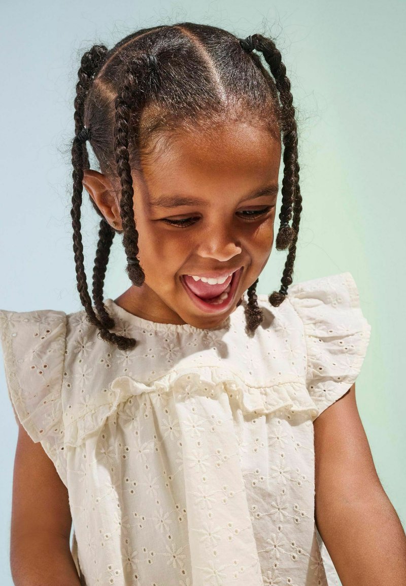 Weißes gesticktes Kleid mit Rüschärmeln, das florale Muster aufweist. Ein Kind mit geflochtenen Haaren lächelt und schaut auf etwas hinunter.