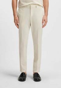 Man som bär beige skräddarsydda byxor och svarta läderskor stående mot en vit bakgrund.