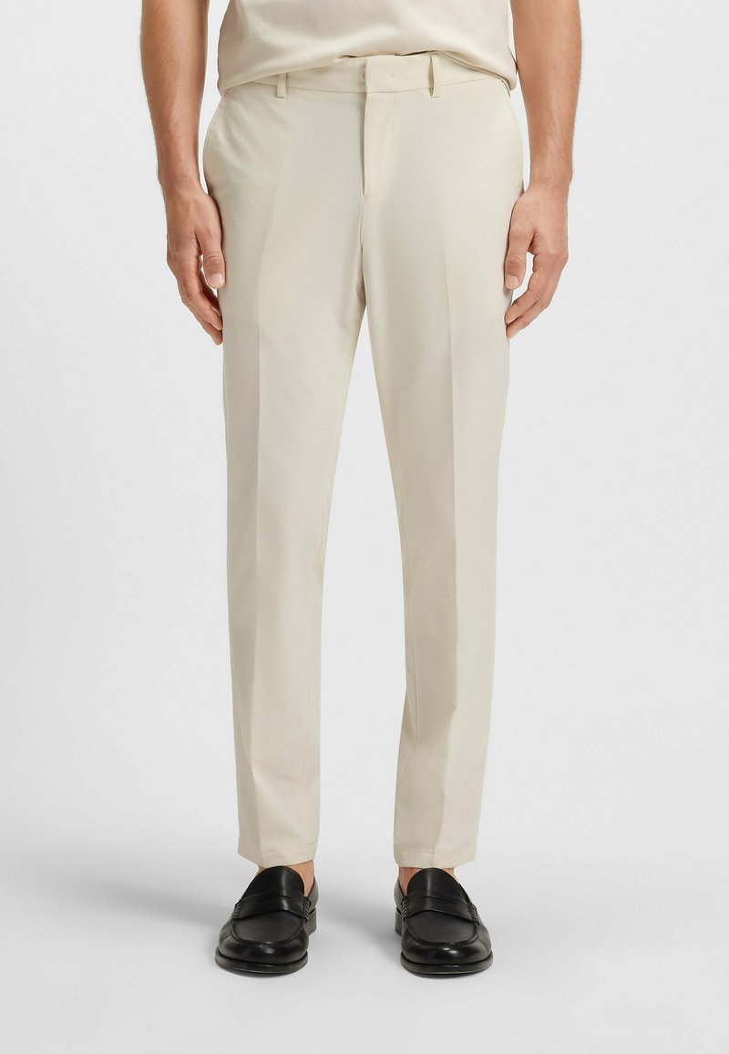 Man som bär beige skräddarsydda byxor och svarta läderskor stående mot en vit bakgrund.