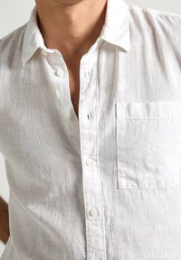 Camicia bianca in lino con abbottonatura frontale, colletto e tasca sul lato sinistro. Il tessuto ha un aspetto strutturato e leggermente trasparente.