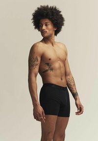 Boxers negros con una cinturilla de marca, usados por una persona sin camiseta y con tatuajes, sobre un fondo neutro.