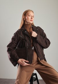 Hnědá semišová kabelka s prohnutými rukojeťmi, vystavená s odpovídající hnědou oversized bundou a rolákem, na neutrálním pozadí.
