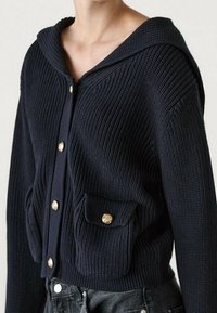 Granatowy sweter z kapturem, wykonany na drutach, z złotymi guzikami, prążkową teksturą oraz kieszenią z przodu. Zaprojektowany w dopasowanej sylwetce.