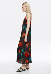 Frau in einem ärmellosen schwarzen Maxikleid mit roten und grünen Blumenmustern, dünnen Schulterträgern und offenen Sandalen mit niedrigen Absätzen.