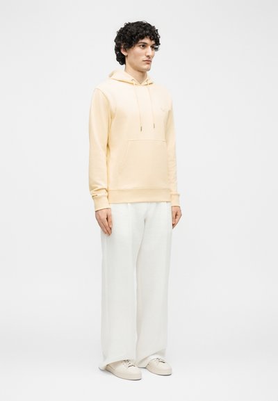 Νέος άνδρας με σγουρά μαλλιά που φοράει μπεζ φούτερ με κουκούλα, λευκό παντελόνι με φαρδιά μπατζάκια και παπούτσια σε ανοιχτό λευκό χρώμα, στέκεται μπροστά σε απλό φόντο.