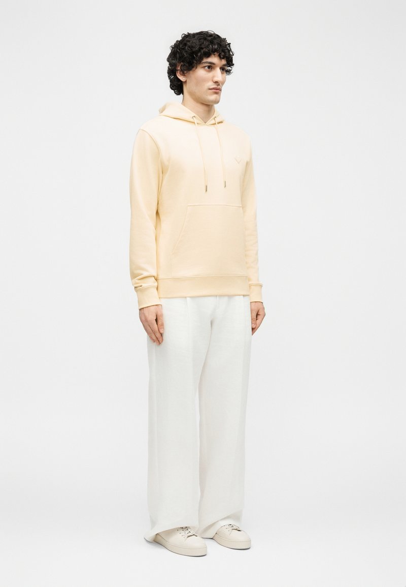 Young man with curly hair wearing a beige hoodie, white wide-leg pants, and off-white sneakers, standing against a plain background.