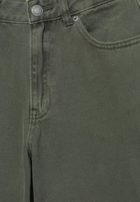 Közeli kép olívazöld farmernadrág elejéről, amely megmutatja az elülső zsebet, övbütyköt, cipzáras szárnyat és a márkajelzéssel ellátott fém gombot.