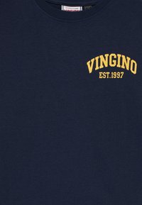 Džinsinio mėlynumo apvalios apykaklės marškiniai su geltonu tekstu "VINGINO EST. 1997" viršutiniame kairiajame krūtinės kampe.
