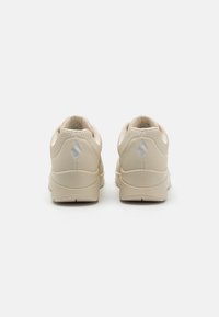 Beige sneakers with a textured finish, rounded heel, and a subtle logo accent on the back. Features padded collar and breathable perforations.