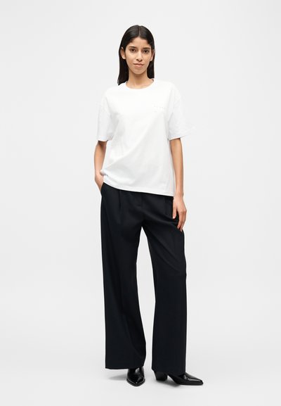 Λευκό βαμβακερό μπλουζάκι με στρογγυλή λαιμόκοψη, κοντά μανίκια, συνδυασμένο με μαύρο παντελόνι φαρδιάς γραμμής με ρίγες και μαύρα μυτερά παπούτσια.