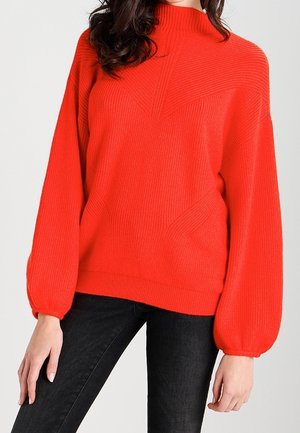 Femme portant un pull tricoté rouge vif avec des manches ballon et un col montant, associé à un pantalon noir ajusté.