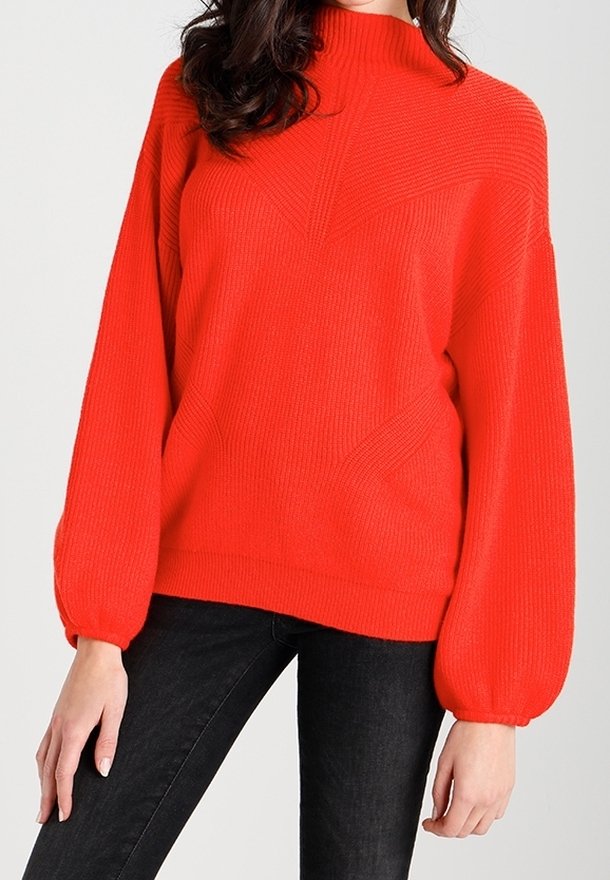 Femme portant un pull tricoté rouge vif avec des manches ballon et un col montant, associé à un pantalon noir ajusté.