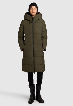 Olive green puffer coat with a hood, featuring a full zipper, side pockets, and a knee-length design. Black textured beanie and tall boots.