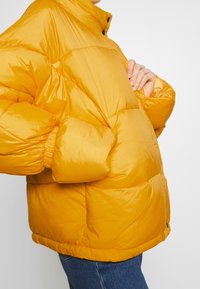 Giacca a vento gialla con finitura lucida, colletto alto, polsini elasticizzati e design corto. Il rivestimento spesso crea una silhouette volumetrica.