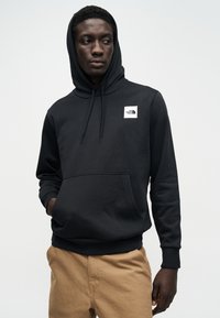 Μαύρο φούτερ με κουκούλα και μπροστινή τσέπη, με μικρό λευκό λογότυπο στο στήθος. Το ύφασμα φαίνεται ομαλό με άνετη εφαρμογή.