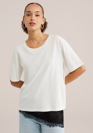 Naine, kellel on suurte rõngakujuliste kõrvarõngastega, kannab lahtist valget T-särki musta pitsist servaga ülevalt ja helerohelisi teksaseid, taust on ühevärviline.