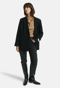 Blazer noir à double boutonnage sur un polo beige, associé à un jean noir et des bottes noires jusqu'aux genoux. Tissu lisse, design structuré.