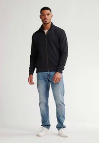 Schwarze Herrenjacke mit Reißverschluss aus weichem Material, mit hohem Kragen und Logo-Akzent, kombiniert mit hellblauen Jeans und weißen Turnschuhen.