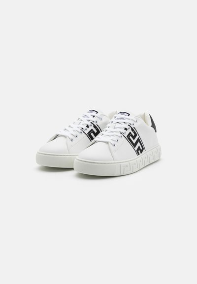 White leather sneakers with black logo detail, round toe, textured rubber sole, and contrasting black heel accent. Laced design.