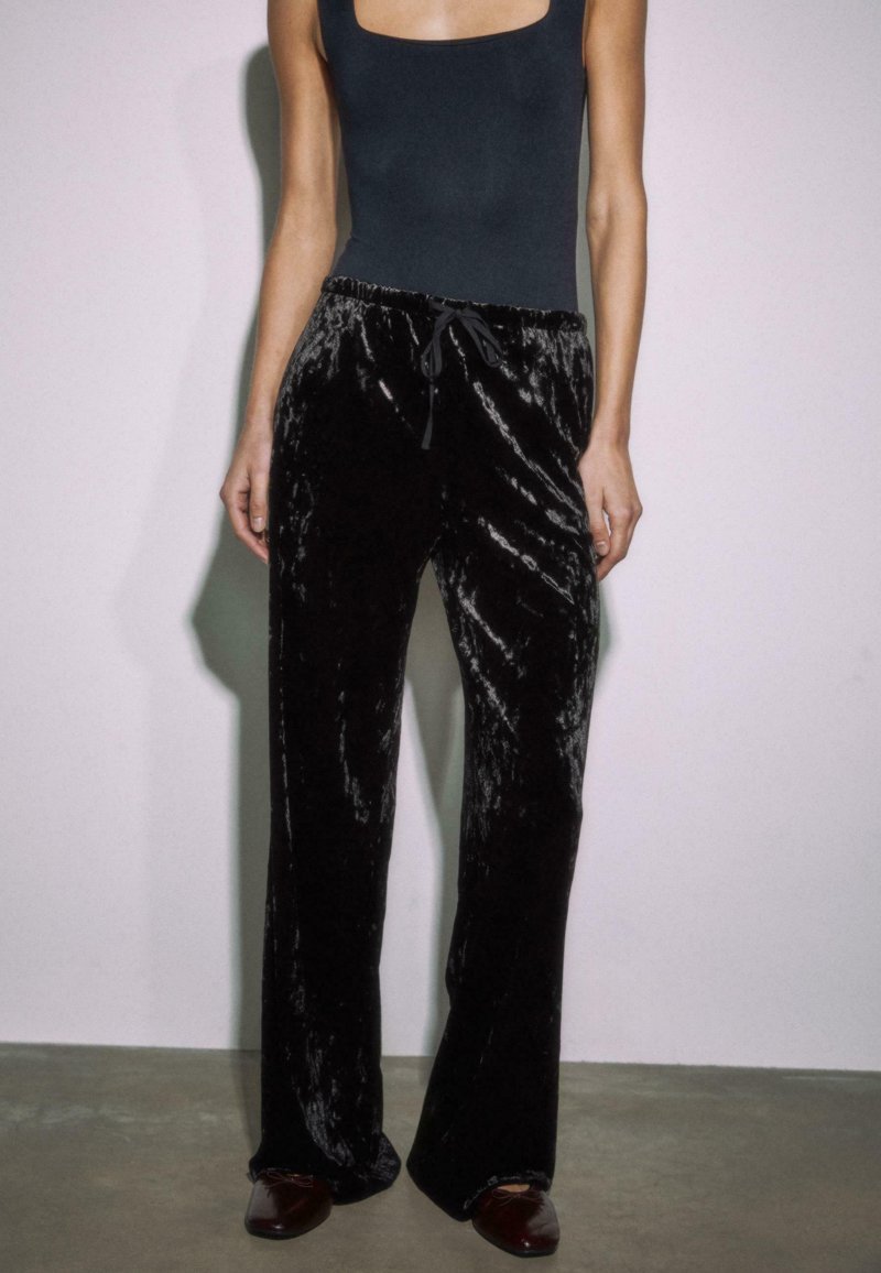 Osoba v čiernych sametových nohaviciach s nastaviteľným pásom, čiernom bezrúškovom topánku a tmavohnedých štvorcových topánkach, stojí pred svetlou jednofarebnou stenou.