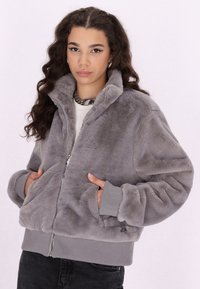 Jachetă din blană sintetică gri, cu fermoar frontal, manșete ribbate și două buzunare laterale. Textură moale și croială relaxată.