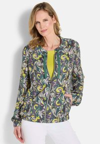 Bedruckte Zip-Jacke mit einem grünen, gelben und lila Wirbelmuster; verfügt über Fronttaschen und einen gerippten Saum. Getragen über einem strukturierten gelben Oberteil.