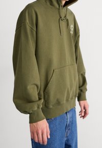 Oliwkowa, hoodie z kapturem, z mieszanki bawełny, w oversize'owym fasonie, z kieszenią typu kangur, regulowanym sznurkiem oraz ściągaczami przy mankietach i dolnym wykończeniu.