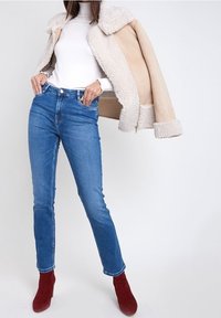 Chaqueta de piel de oveja beige con cuello alto, suéter ajustado blanco de cuello de tortuga, jeans de mezclilla azul y botas de tobillo rojas.