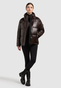 Bruine gewatteerde jas met een glanzende afwerking, hoge kraag, knopen aan de voorkant, zijzakken, gedragen over een zwarte fitted turtleneck en leggings. Zwarte laarzen.