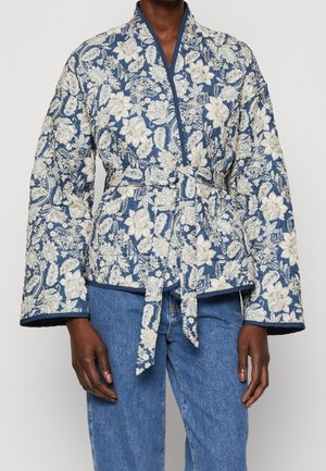 Persoon draagt een blauw-wit gebloemd gewatteerd jasje met ceintuur in de taille en wijde mouwen, gecombineerd met een blauwe spijkerbroek.