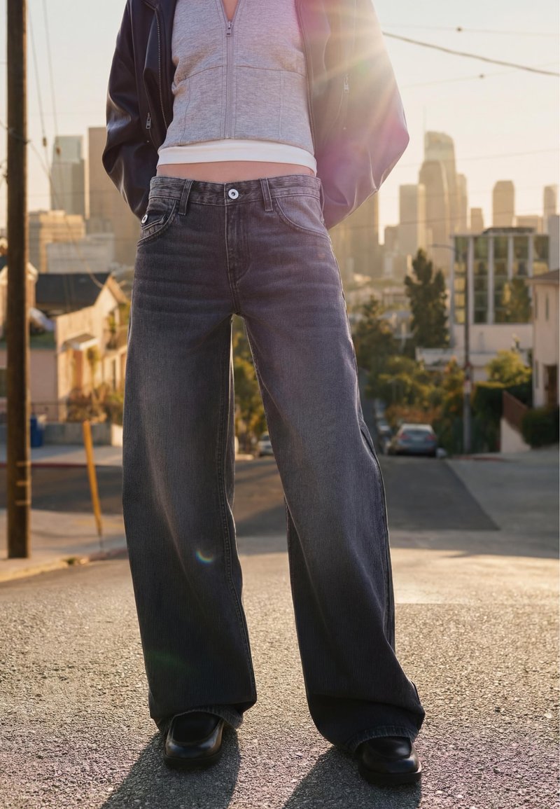 Personne portant un jean large foncé, des chaussures noires et un sweat à capuche gris coupé, debout dans une rue de la ville avec le soleil et des bâtiments en arrière-plan.